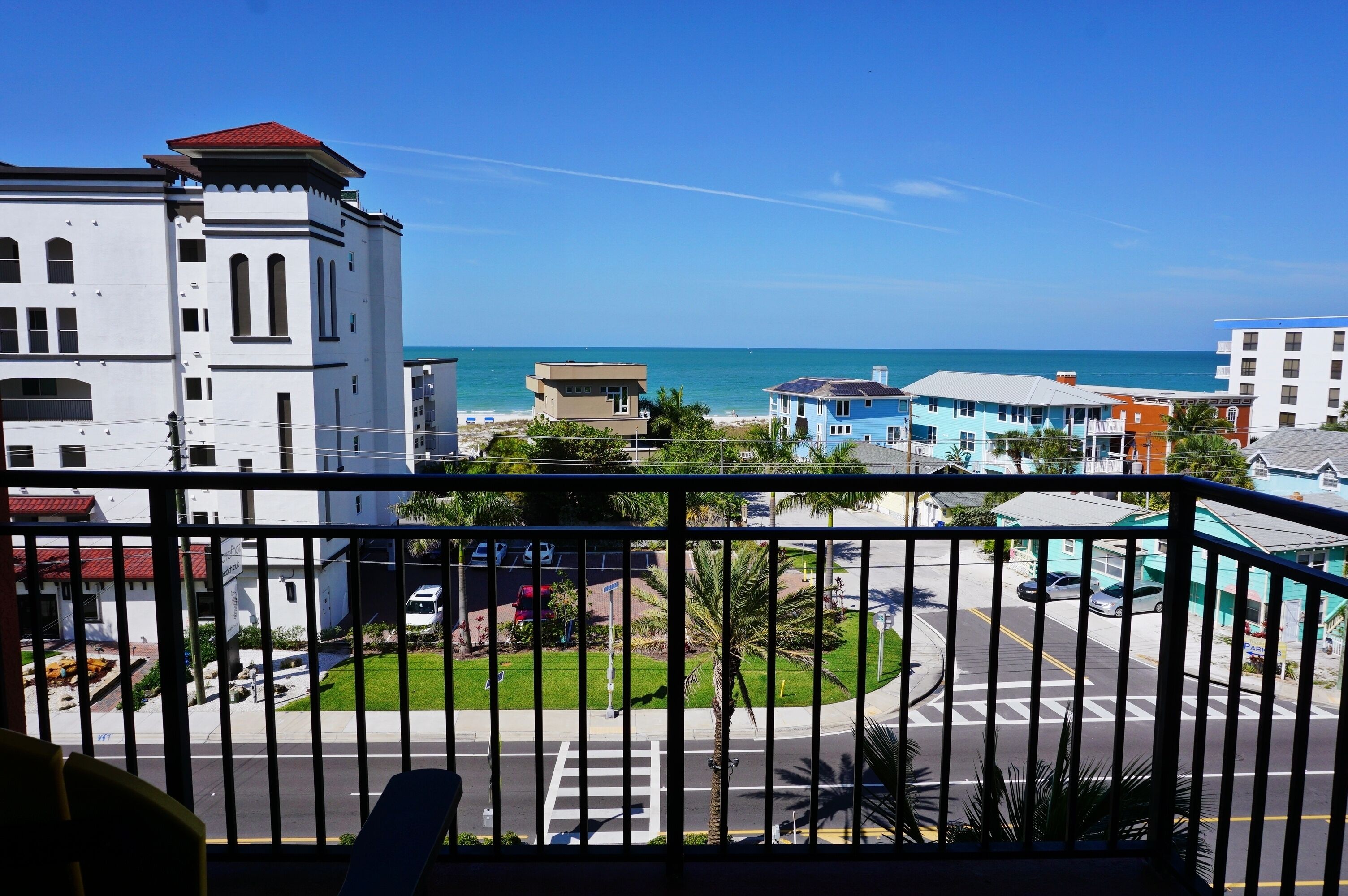 Vista costeira a partir de uma varanda, com céu azul límpido, um edifício branco com janelas em arco e uma estrada com palmeiras.