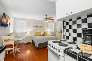 A compact kitchen with a stove, a dining area with a table and chairs, a bed with a headboard, and a ceiling fan.