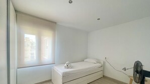 A small, single-bed room with a wooden floor, a window with blinds, a bedside table, and a standing fan.