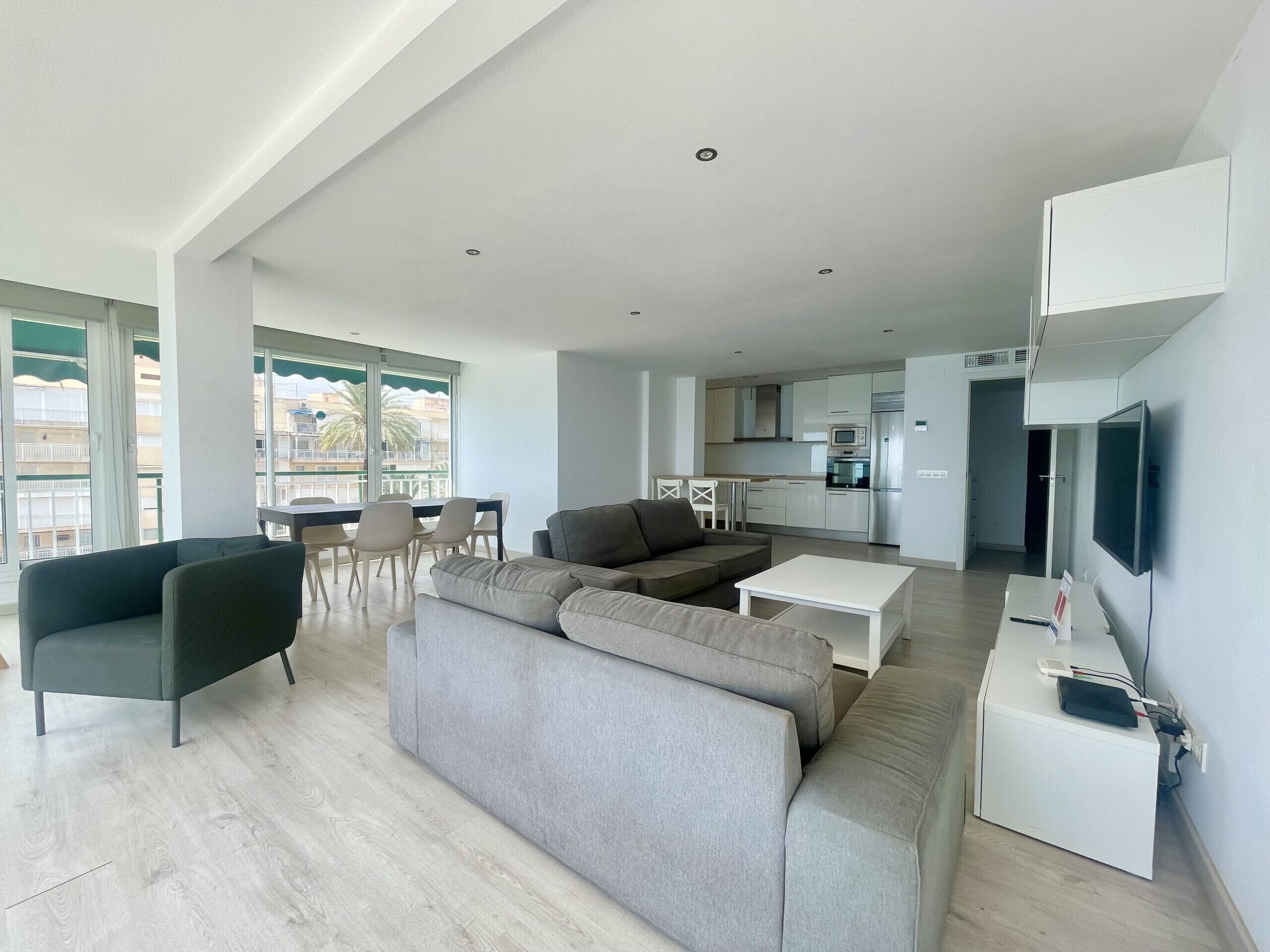 A modern living room with a grey sofa, a dining area with chairs, and a kitchen in the background.