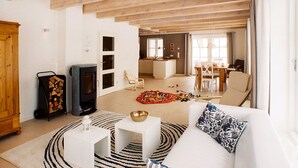 Een ruime woonkamer met een witte bank, een salontafel en een open haard. Achtergrond toont een eethoek met een tafel en stoelen.