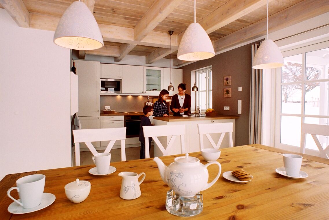 Une cuisine moderne avec des meubles blancs, une table à manger en bois et de la vaisselle blanche, ainsi que des luminaires suspendus.