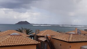A coastal view with terracotta-roofed buildings, a mountain in the distance, and the ocean with waves. - SEAHAUS FUERTE in a residential complex directly on the sea. (Corralejo)