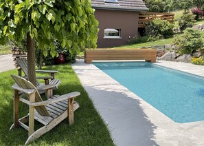 A swimming pool with a wooden deck, a bench, and a tree with green leaves. - Cottage in the forest with heated pool from May to October (Buhl)