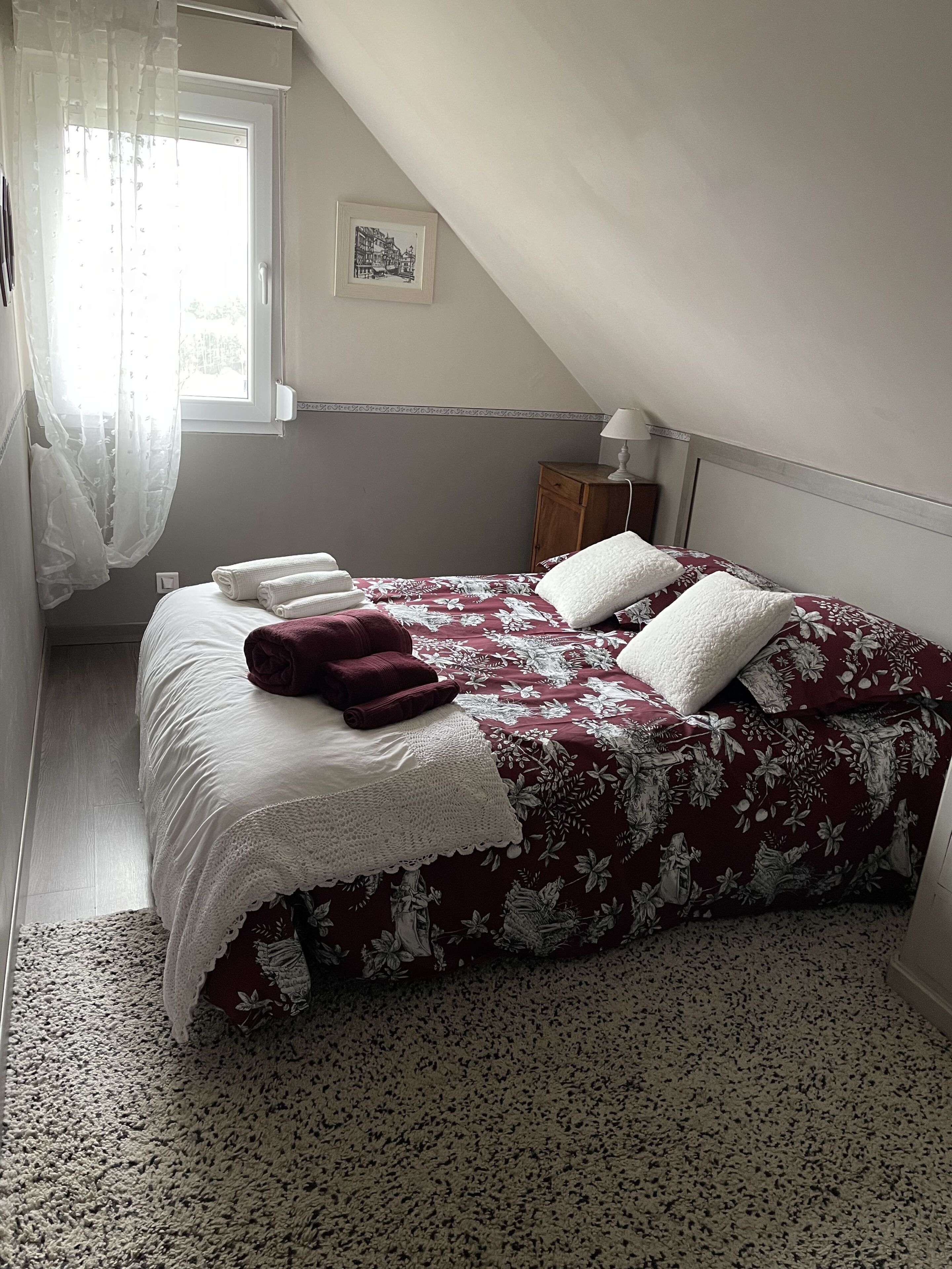 Un dormitorio con cama, ventana con cortinas de gasa, un cuadro enmarcado en la pared y una mesita de noche de madera con lámpara.