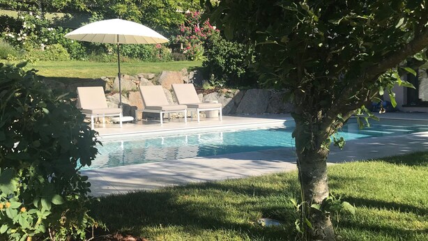 A swimming pool with lounge chairs and an umbrella, surrounded by greenery and a stone wall. - Cottage in the forest with heated pool from May to October (Buhl)