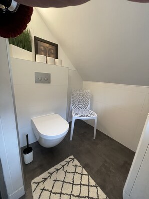 A small, modern bathroom with a white toilet, a white chair, and a floor mat. - Cottage in the forest with heated pool from May to October (Buhl)