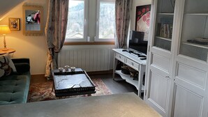 A cozy room with a loft bed, a small table with a lamp, a TV on a white cabinet, and a window with curtains. - Cottage in the forest with heated pool from May to October (Buhl)