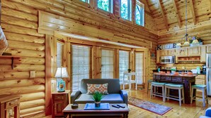 A log cabin interior with a living area, sofa, coffee table, kitchen, and large windows.