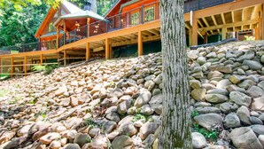 A cabin with a wooden deck, stone foundation, and a large tree in the foreground.
