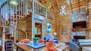 A wooden cabin interior with a dining area, a living room, a fireplace, and a staircase.
