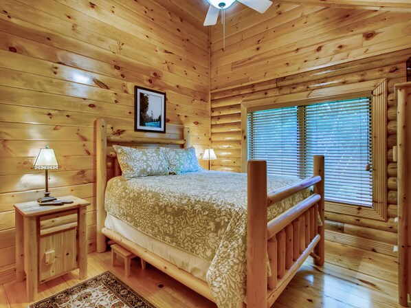 Un dormitorio de cabaña de madera con una cama, mesitas de noche, un ventilador de techo y una ventana con persianas.
