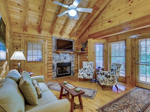 A cozy log cabin interior with a fireplace, a sofa, a television, and a ceiling fan.