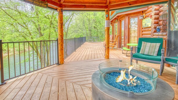 A wooden deck with a glass fire pit, outdoor seating, and a view of the surrounding forest.