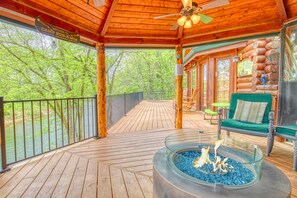 Una terraza de madera con un brasero de vidrio, mobiliario exterior y vistas al bosque circundante.