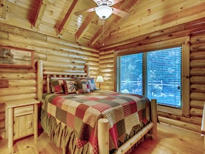 A log cabin bedroom with a wooden ceiling, a bed with a quilt, a nightstand, and a window with blinds.