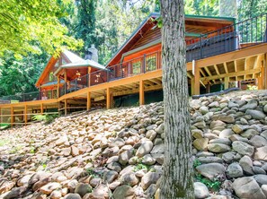 A cabin with a wooden deck, stone foundation, and a large tree in the foreground.