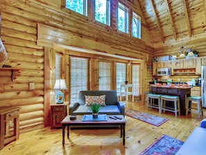 A log cabin interior with a living area, sofa, coffee table, kitchen, and large windows.