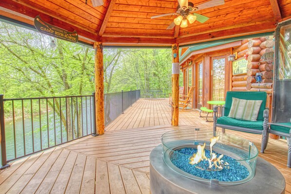 A wooden deck with a glass fire pit, outdoor seating, and a view of the surrounding forest.