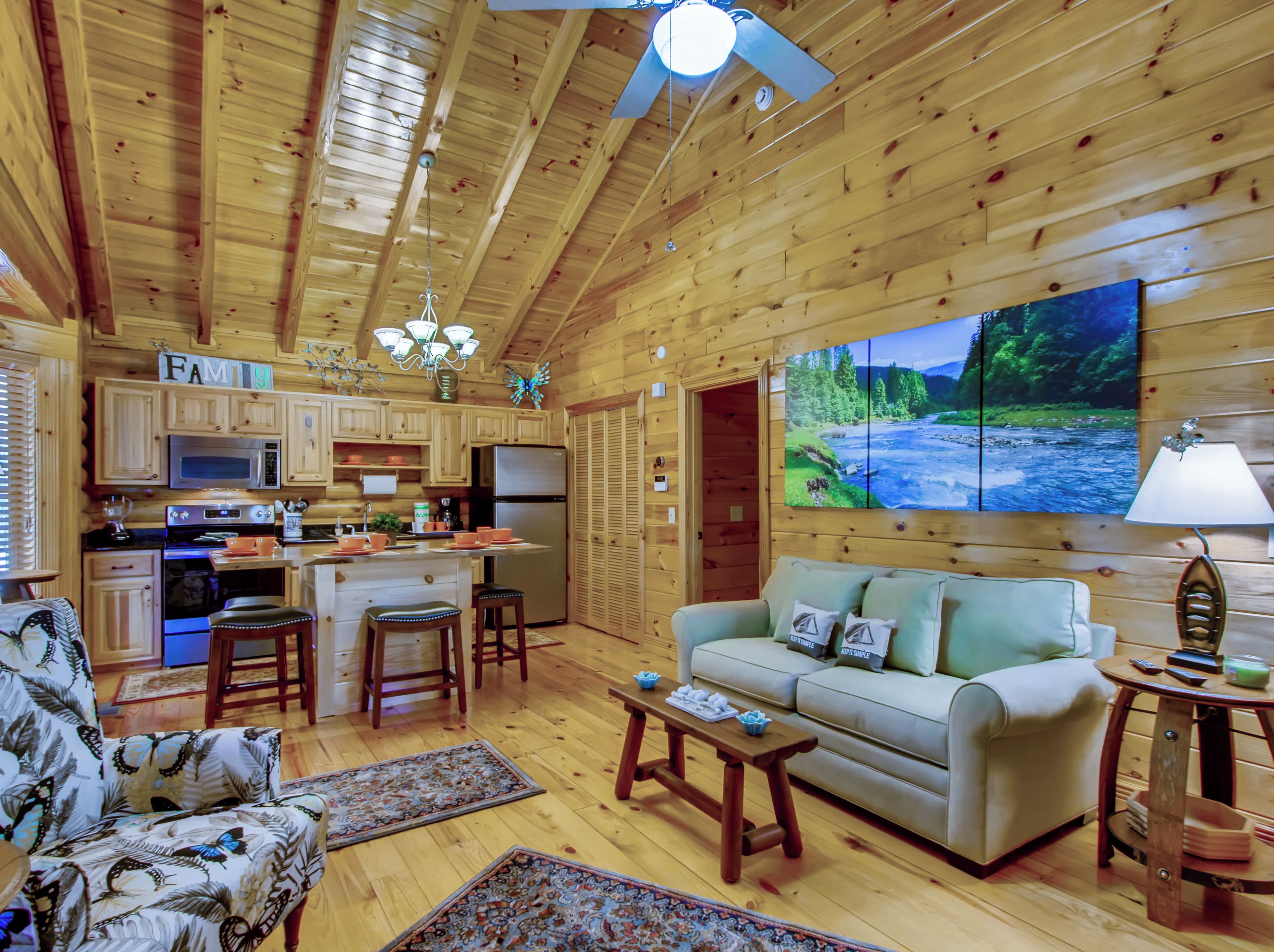 Ein gemütliches Blockhaus-Interieur mit Holzwänden, einer Küche mit modernen Geräten, einem bequemen Sofa und einem großen Fernseher, der eine Naturansicht zeigt.