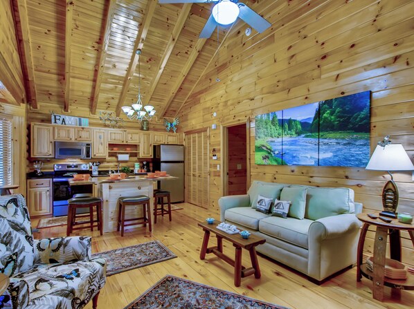 Interior acogedor de una cabaña con paredes de madera, una cocina con electrodomésticos modernos, un sofá cómodo y un televisor grande que muestra una escena de la naturaleza.