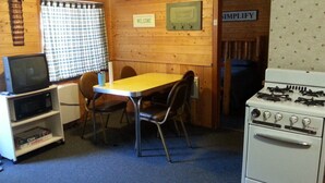 Une petite pièce agréable et chaleureuse, avec un intérieur en bois, une table à manger entourée de chaises, un téléviseur posé sur une étagère et un poêle.
