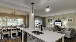 Eine moderne Küche mit Kochinsel, Essbereich mit weißen Stühlen und ein Wohnzimmer mit Sofa und Fernseher.