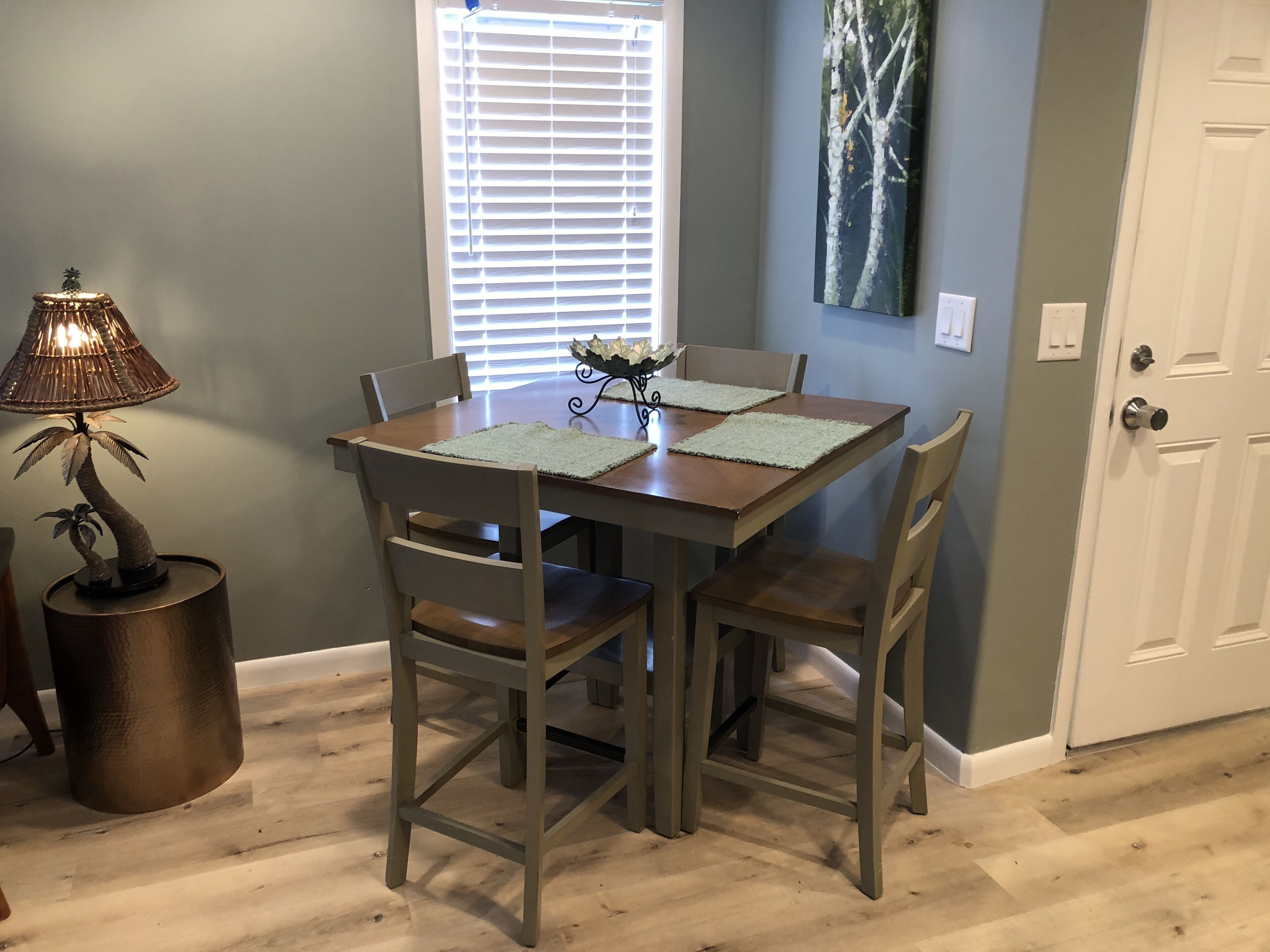 Un comedor con una mesa de madera, cuatro sillas, una lámpara decorativa y una ventana con persianas.