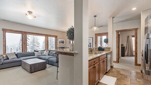 Een ruime woonkamer met een grijze modulaire bank, een houten salontafel en een groot raam met uitzicht op besneeuwde bomen.