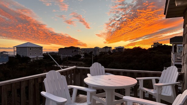 Terraza de madera con sillas Adirondack blancas y una mesa redonda, con vistas a una zona residencial al atardecer.