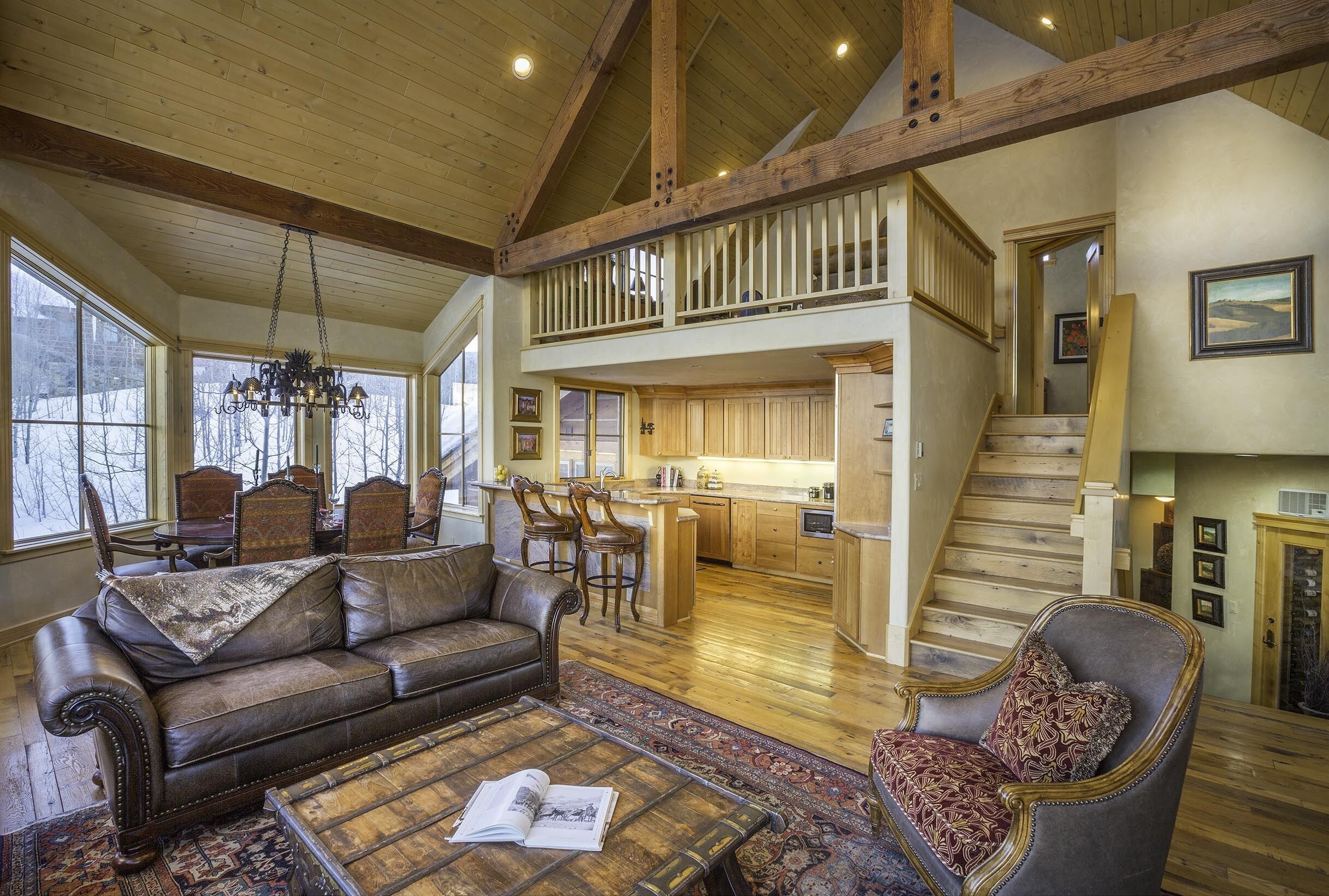 A spacious living area with a leather sofa, a patterned armchair, and a coffee table with a book. A wooden staircase leads to an upper level with a balcony.