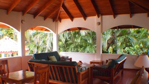 A covered outdoor seating area with wooden furniture and large windows overlooking a tropical garden.