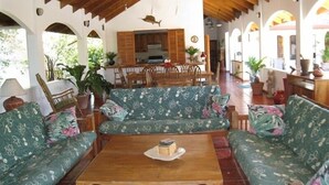A spacious living area with a wooden ceiling, a central sofa set, a coffee table, and a kitchen in the background.