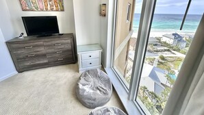 A bedroom with a large window overlooking a beach, a TV on a dresser, and a small bedside table. - Beautiful Sunset paradise with New Golf Cart 4811 (Miramar Beach)