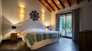 Un dormitorio con una cama grande, vigas de madera en el techo, dos lámparas de pared y vistas al exterior a través de un ventanal corredero.
