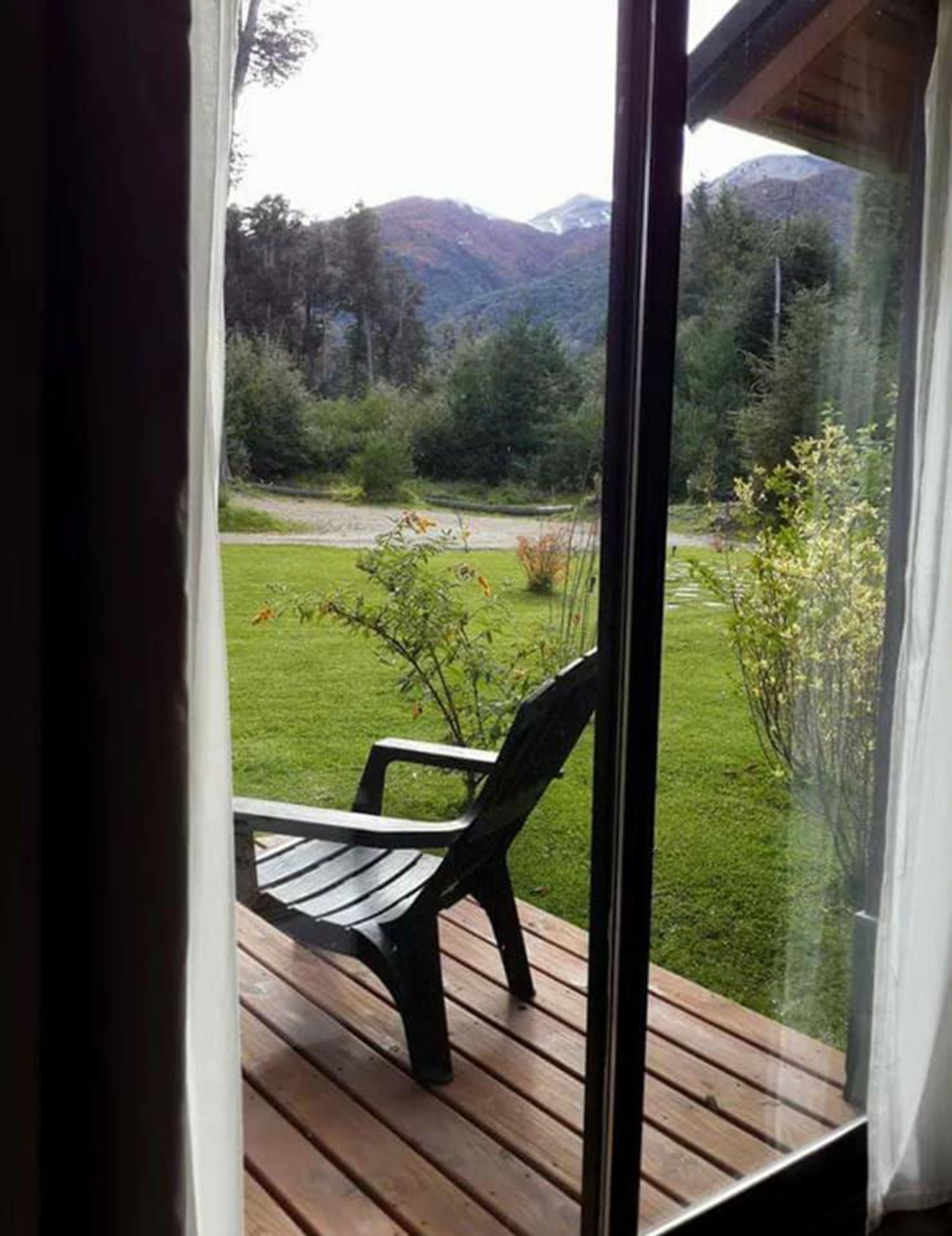 Vista desde una ventana con terraza de madera y una silla, con vistas a un jardín y montañas.