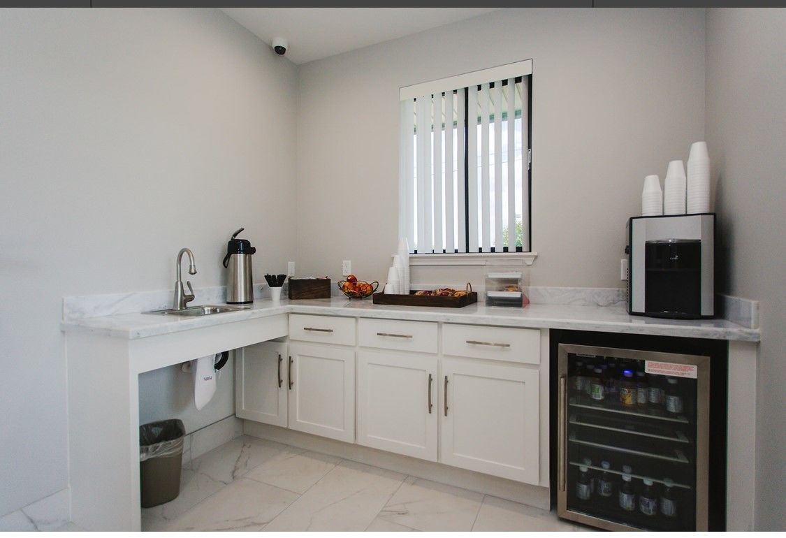 A modern kitchen with white cabinets, a stainless steel sink, a window with blinds, and a refrigerator with a water dispenser.