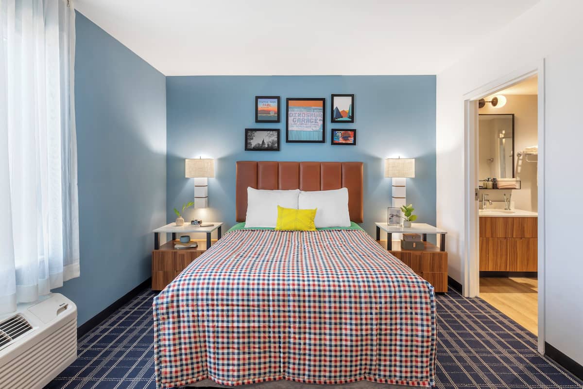 A hotel room with a checkered bedspread, a headboard, bedside lamps, a nightstand, a window with curtains, and a wall with framed pictures.