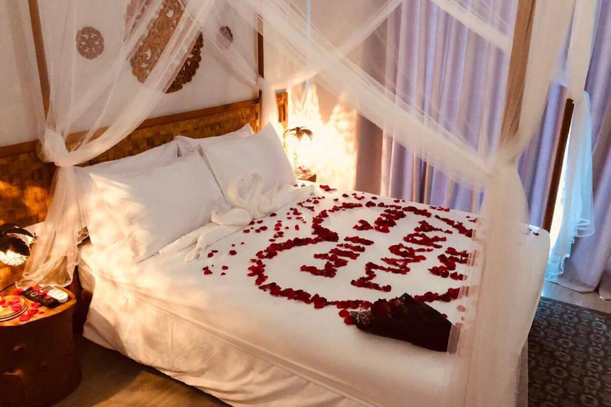 A bed with a canopy, rose petals on the bed, and a bedside table with a phone and a bottle.