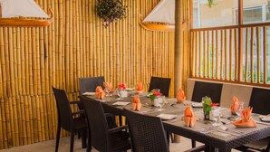 Un espace repas agrémenté d’un mur en bambou, de chaises en osier noir et de serviettes orange.