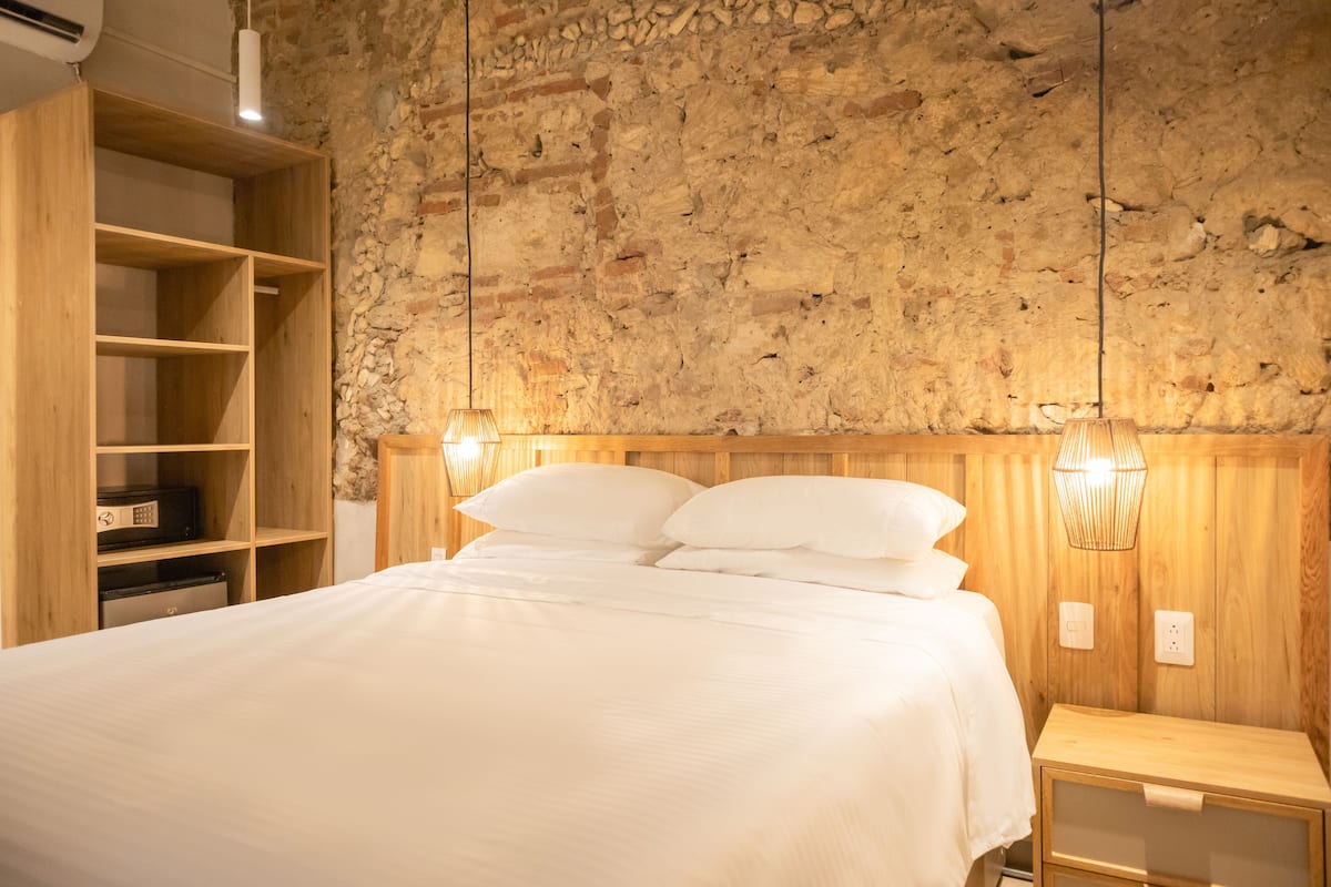 Een slaapkamer met een stenen muur, een houten bed en witte beddengoed, en een nachtkastje met een lamp.