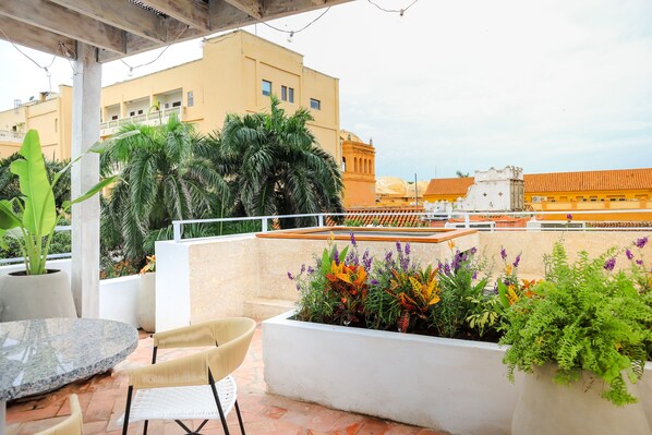 A rooftop terrace with a round table and chairs, surrounded by potted plants and a view of buildings in the background.