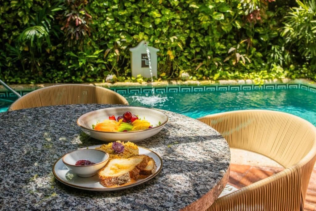 Una mesa redonda con platos de comida y un tazón pequeño, rodeada de sillas de mimbre, con una piscina y abundante vegetación al fondo.