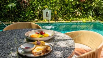 Una mesa redonda con platos de comida y un tazón pequeño, rodeada de sillas de mimbre, con una piscina y abundante vegetación al fondo.
