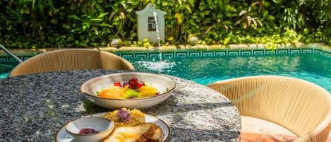 Ein runder Tisch mit Speisen und einer kleinen Schüssel, umgeben von Korbstühlen, mit einem Pool und üppigem Grün im Hintergrund.