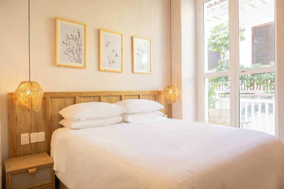 Een net opgemaakt bed met witte lakens, een houten hoofdbord en twee wandlampen. Daarboven hangen drie ingelijste kunstwerken.