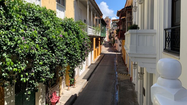 Une rue étroite, bordée de bâtiments des deux côtés, un ciel dégagé et une petite flaque d’eau sur le sol.