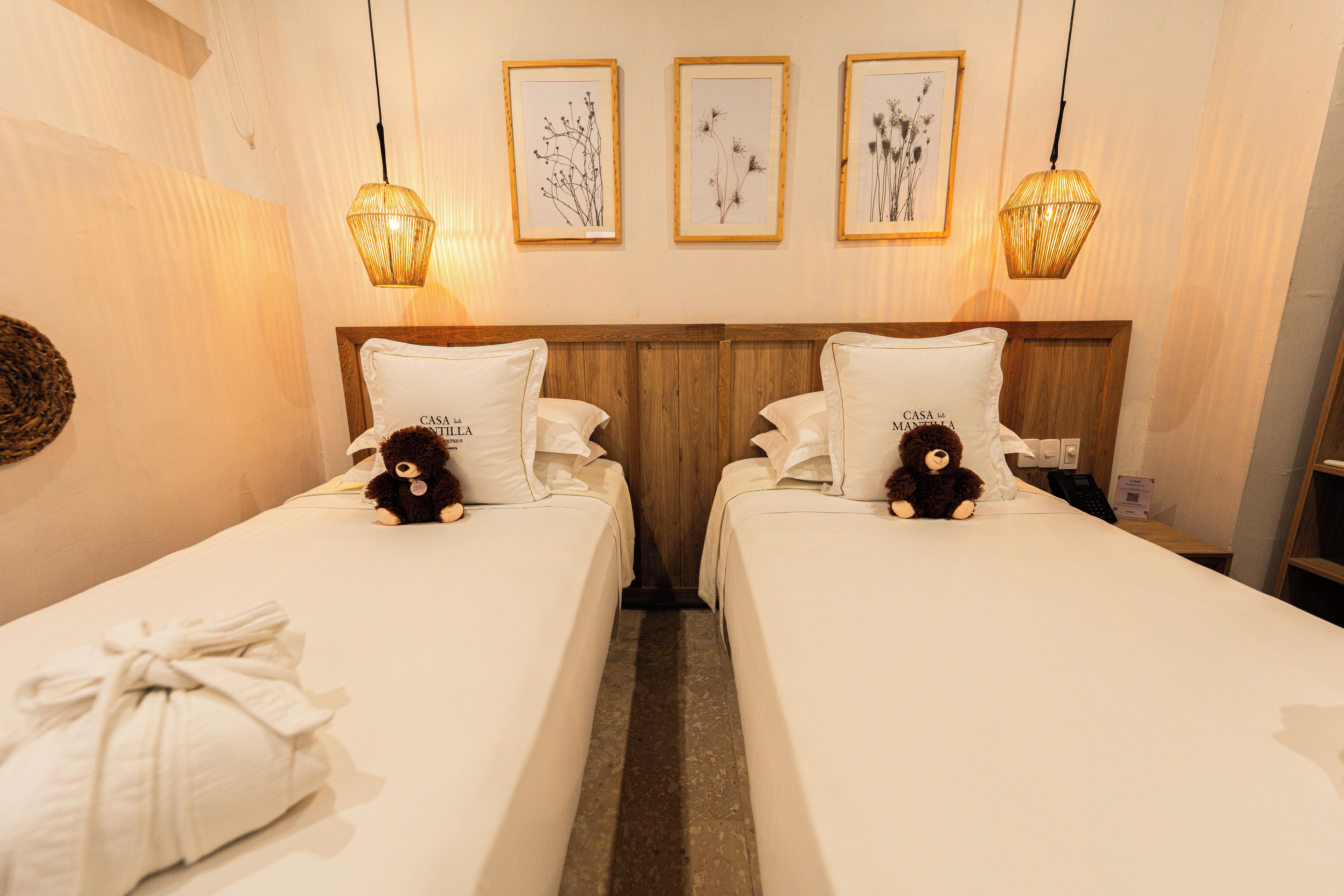 A hotel room with two beds, each with a pillow labeled 'CASA LA MANTILLA,' a teddy bear, and a towel on the left bed. There are three framed pictures on the wall above the beds.