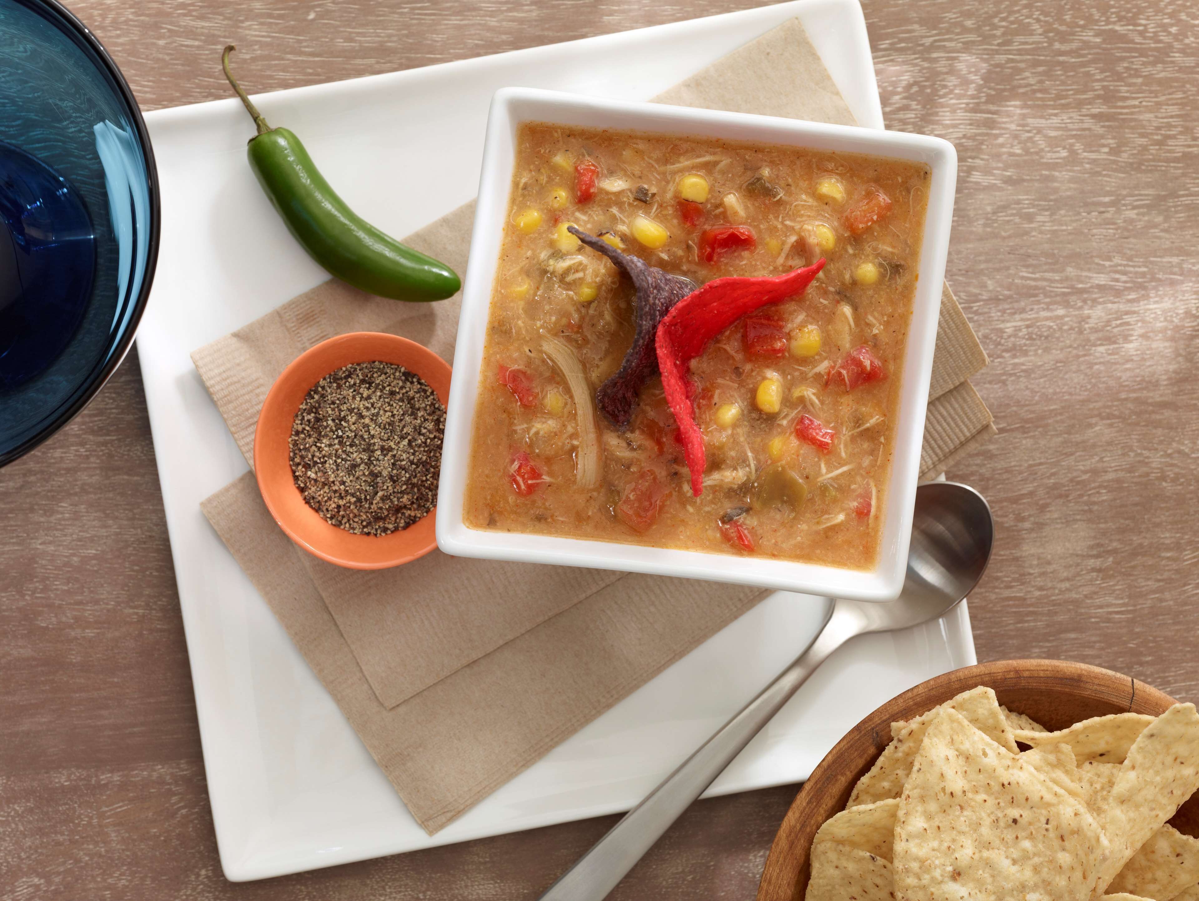 Un tazón de sopa con un chile rojo encima, un tazón pequeño de pimienta negra y un tazón de totopos sobre una mesa de madera.