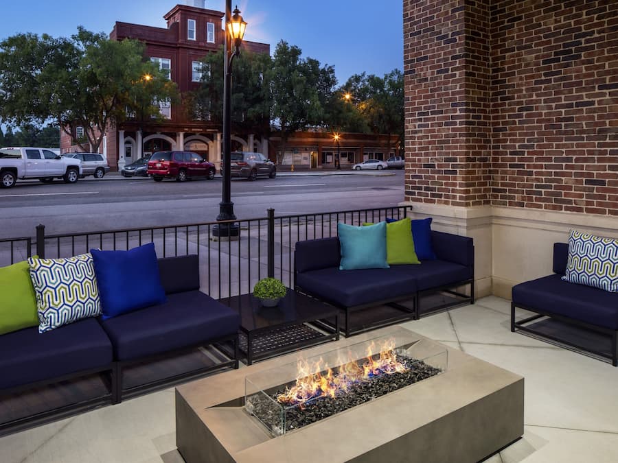 An outdoor seating area with a fire feature, a brick building, and parked cars in the background.